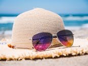 Close-up of straw hat and sunglasses on a sandy beach with ocean waves in the background.